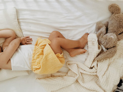 A toddler sleeping in bed, kicking the blanket off of her with her feet.